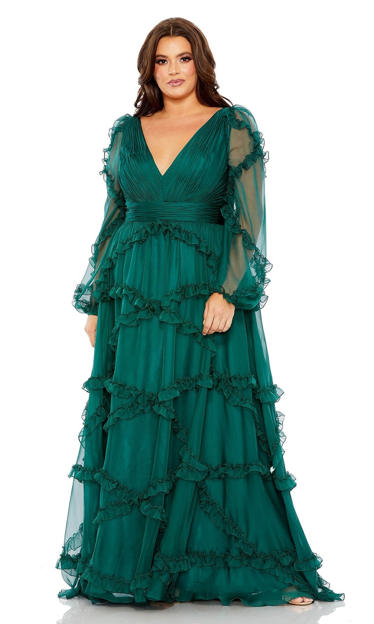 A woman models the Long Plus-Size Prom Dress 68425 by Mac Duggal—an emerald green gown with a v-neckline, sheer long sleeves, and tiered ruffled layers—posing against a white background.